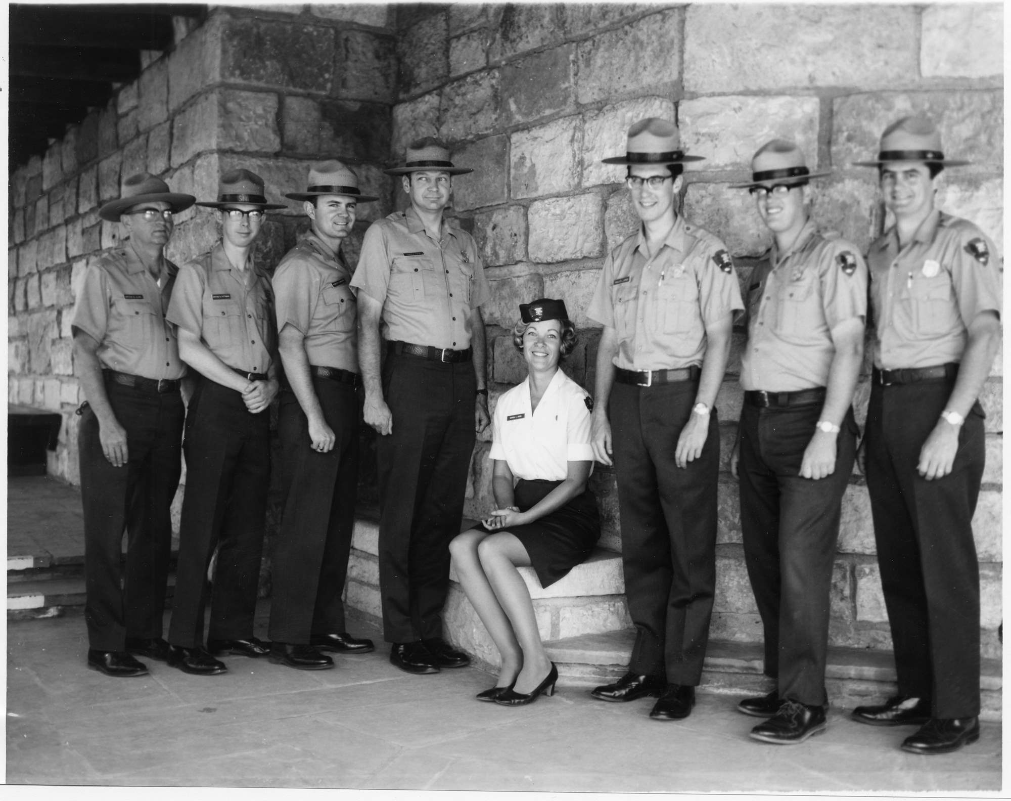 Marion Riggs in NPS uniform skirt, blouse, small arrowhead pin, and pillbox hat sits between seven men standing in uniforms with shield-shaped badges.